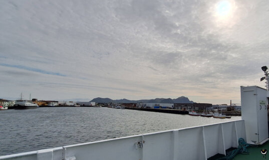 Blick vom Deck der Fähre auf den Hafen von Andenes mit Fischerbooten, Hafenanlagen und Bergen im Hintergrund.