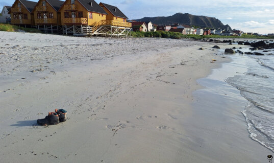 Heller Sandstrand von Bleik mit Holzhäusern auf Stelzen, kleinen Wellen am Ufer und der markanten Bergkulisse im Hintergrund.