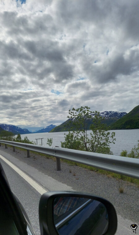 Blick aus dem Auto auf einen norwegischen Fjord mit bewaldeten Berghängen, schneebedeckten Gipfeln und tief hängenden Wolken.