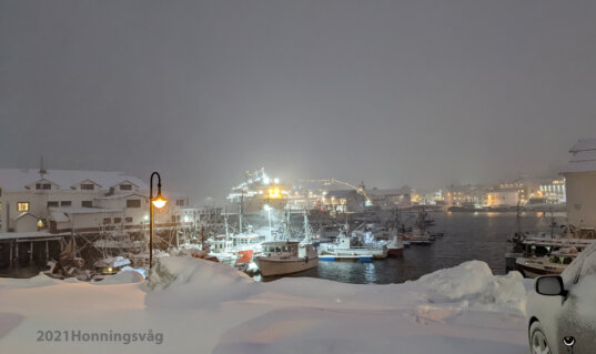 Verschneiter Hafen von Honningsvåg im Winter 2021 mit beleuchteten Fischerbooten und Schiffen.
