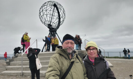 Zwei Reisende vor dem großen Globus am Nordkapp bei bedecktem Himmel.